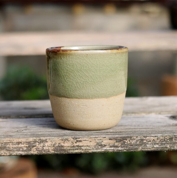 Un « Mug sans anse en grès - émail vert » présente une glaçure craquelée verte sur le dessus et une base beige lisse, reposant élégamment sur une surface en bois. - Atelier Terre et Bois à Colmar