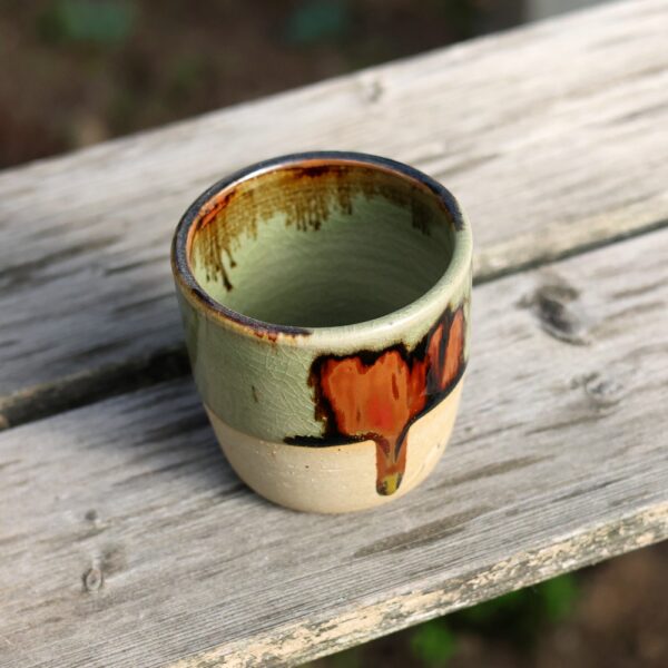 Un « Mug sans anse en grès - émail vert » présentant un superbe jeu de motifs de gouttes marron et orange orne élégamment une surface en bois patiné, illustrant un savoir-faire artisanal exquis qui incarne l'essence de l'art de la céramique. - Atelier Terre et Bois à Colmar