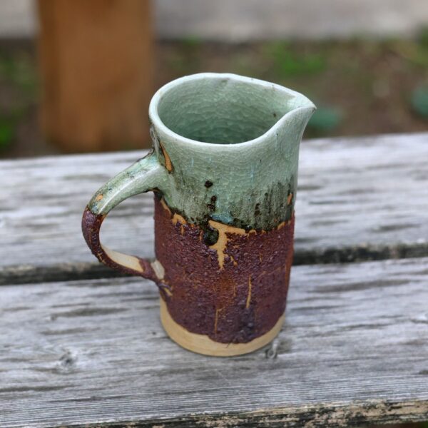 Le "Pichet en grès - terre de récolte ref 7", illustrant un savoir-faire artisanal, présente une base marron terreuse et un plateau vert, reposant élégamment sur une surface en bois patiné. - Atelier Terre et Bois à Colmar