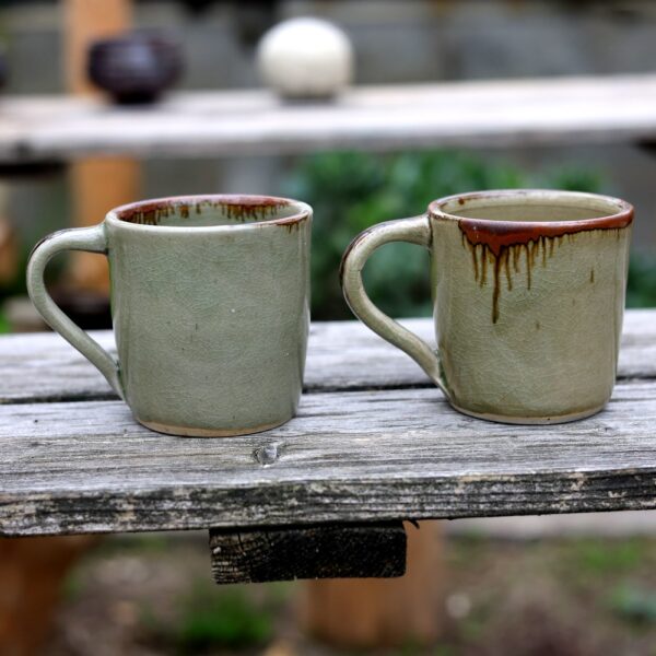 Une paire de mugs en céramique à glaçure verte et à anse gracieuse en grès repose sur une table en bois patiné à l'extérieur, incarnant l'élégance rustique. Lot de 2 Mugs avec anse en grès - réf 112 capture parfaitement ce moment serein. - Atelier Terre et Bois à Colmar