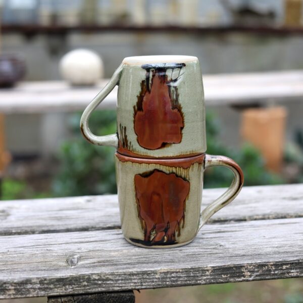 Le Lot de 2 Mugs avec anse en grès - réf 112, présentant des coulures abstraites brunes et oranges, sont empilés à l'envers sur une surface en bois à l'extérieur. - Atelier Terre et Bois à Colmar