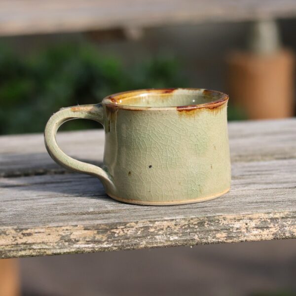 Une tasse rustique en céramique vert clair « tasse avec anse - grès - réf 6 », dotée d'un émail craquelé qui met en valeur son art exquis en céramique, repose gracieusement sur une surface en bois à l'extérieur. - Atelier Terre et Bois à Colmar