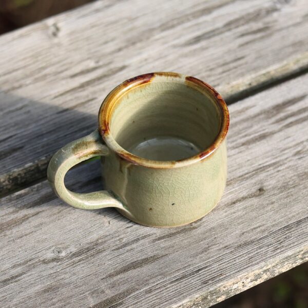 La "tasse avec anse - grès - réf 6", une tasse en céramique verte avec une glaçure craquelée qui met en valeur un savoir-faire exquis, est posée sur une table en bois patiné. - Atelier Terre et Bois à Colmar