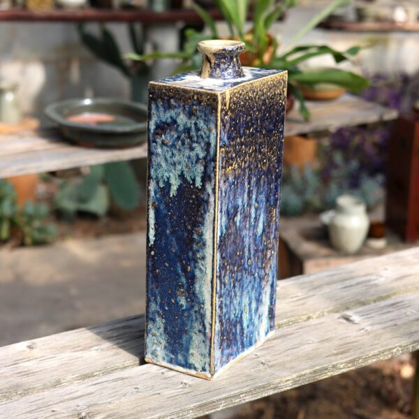 Le Grand vase rectangulaire en grès - Blanc - Bleu - réf 4, de forme haute et émaillée aux tons bleus, est posé sur un banc en bois au milieu d'un décor de jardin. - Atelier Terre et Bois à Colmar