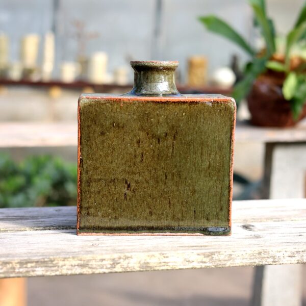 Un vase rectangulaire en grès rouge émaillé de vert (réf. 7) trône gracieusement sur une étagère en bois, incarnant l'esprit de l'artisanat. Des plantes floues et des bougies créent une toile de fond élégante, faisant écho au charme intemporel de la poterie. - Atelier Terre et Bois à Colmar