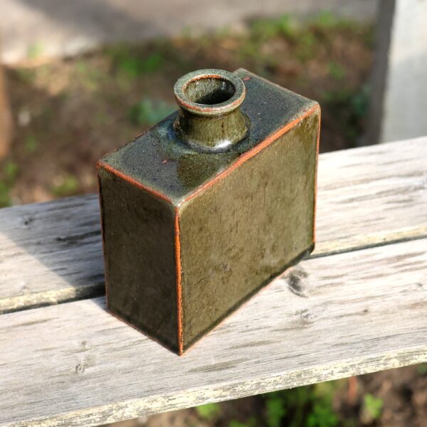 Un vase rectangulaire en grès rouge - émail vert - réf 7, présentant un artisanat exquis avec une ouverture circulaire étroite, est placé sur une surface en bois à l'extérieur. - Atelier Terre et Bois à Colmar