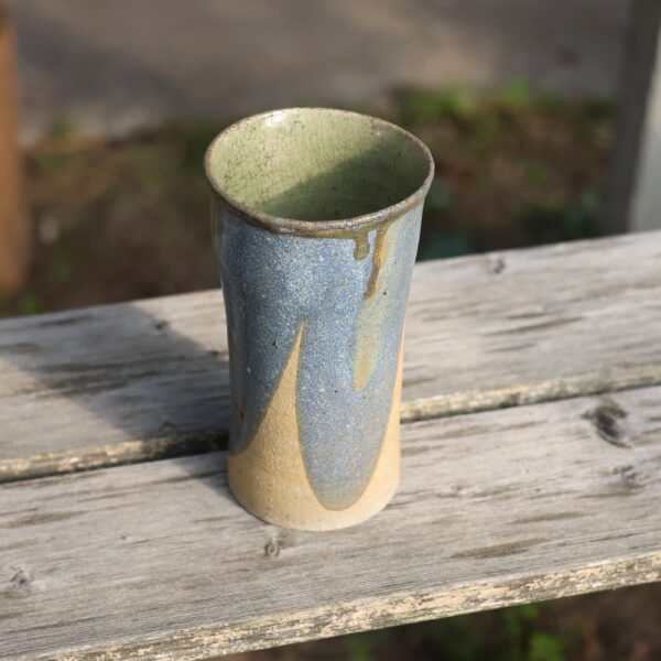 Un vase en céramique nommé « Vase rond en grès ref 56 », doté d'un intérieur vert et d'un extérieur bleu et beige qui met en valeur un savoir-faire exquis, repose sur un banc en bois patiné. - Atelier Terre et Bois à Colmar