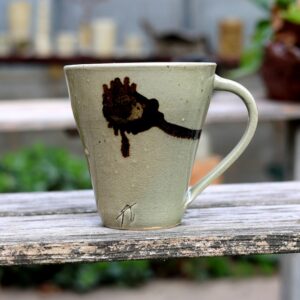 Un grand Mug en grès avec anse - réf 45 au motif floral repose élégamment sur une surface en bois, magnifiquement encadré par un décor extérieur en arrière-plan, capturant véritablement son essence. - Atelier Poterie Terre et Bois à Colmar